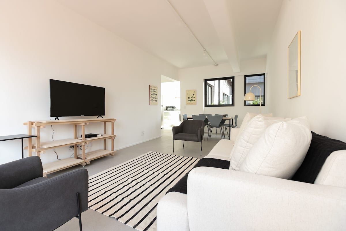 A modern living room with a flat-screen TV, a wooden entertainment unit, a white sofa, a black and white striped rug, and a dining area with chairs and a table.