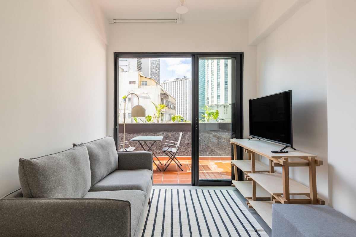 A modern living room with a grey sofa, a flat-screen TV on a wooden stand, a small table, and a sliding glass door leading to an outdoor patio with a table and chairs.