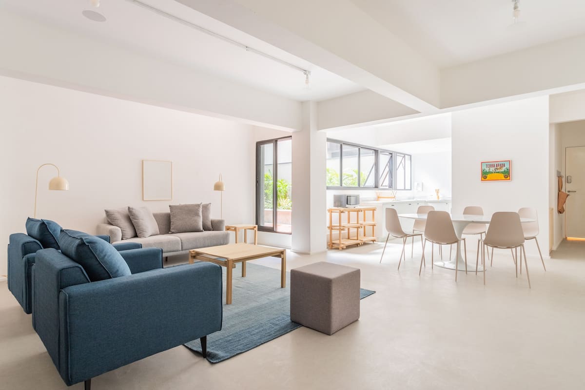 A modern living room with a blue sofa, a wooden coffee table, and a grey ottoman. There is a dining area with a white table and chairs.