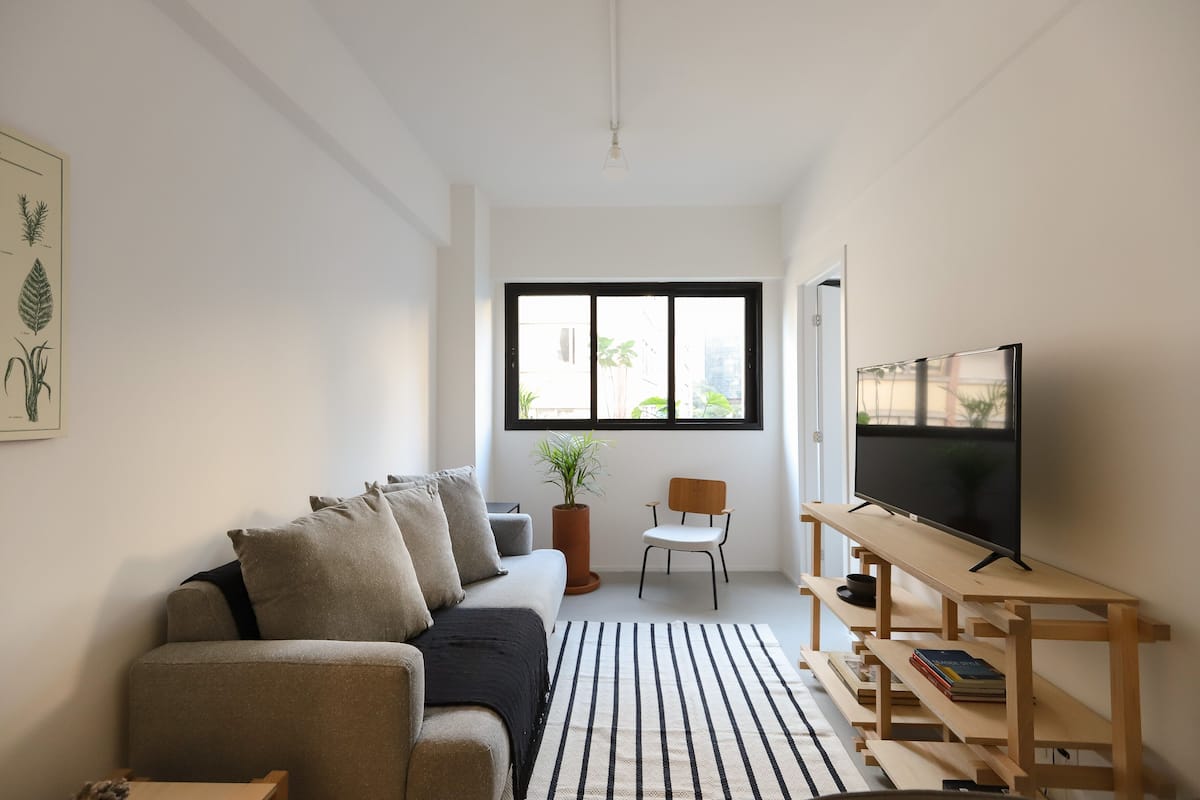 A modern living room with a sofa, a TV on a wooden stand, a potted plant, and a window with a view of buildings.