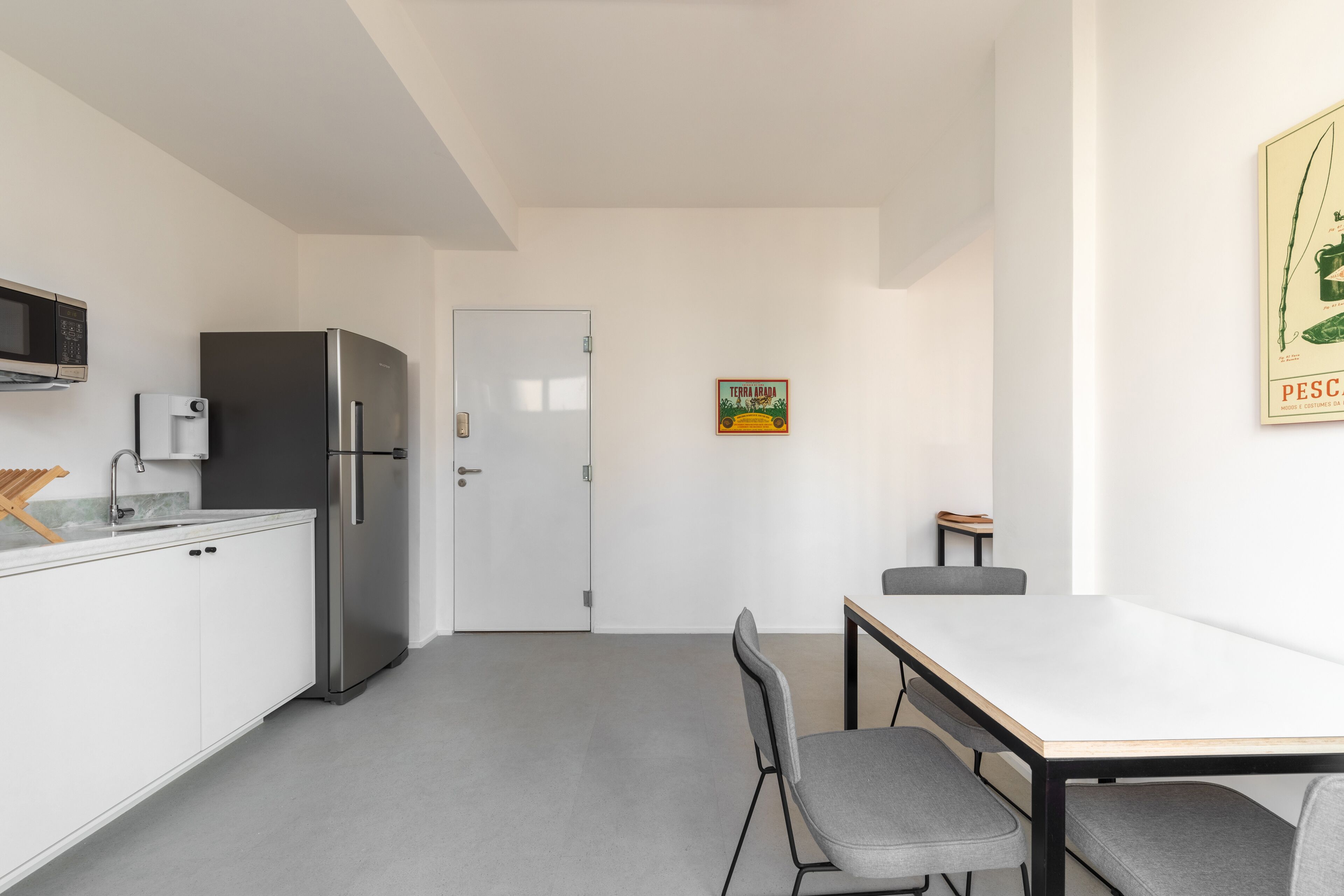 Cozinha moderna com frigorífico, micro-ondas e pia. Há uma mesa de jantar com cadeiras e uma pequena mesa com uma cadeira. Uma porta é visível ao fundo.