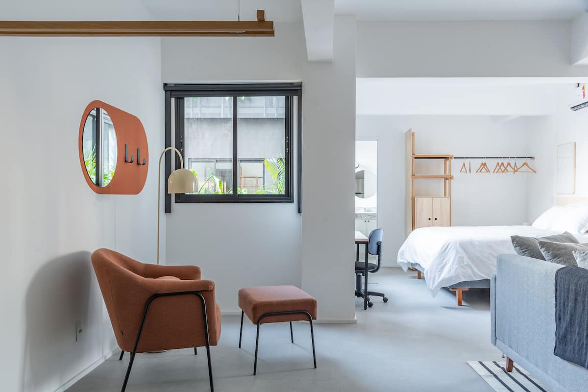 A modern bedroom with a bed, a desk, and a chair. There is a window with blinds, a mirror, and a hanging light fixture.