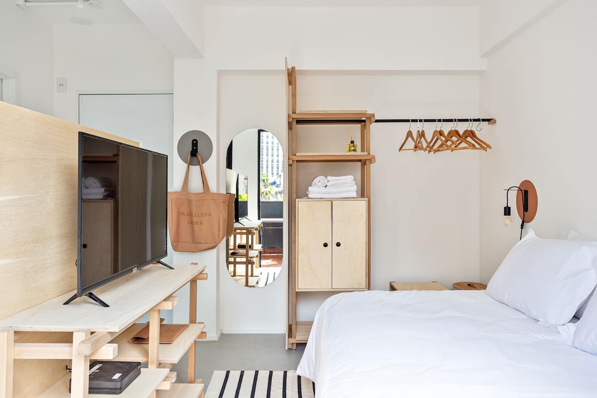 A modern hotel room with a bed, a TV, a wooden desk, a wardrobe, and a mirror.
