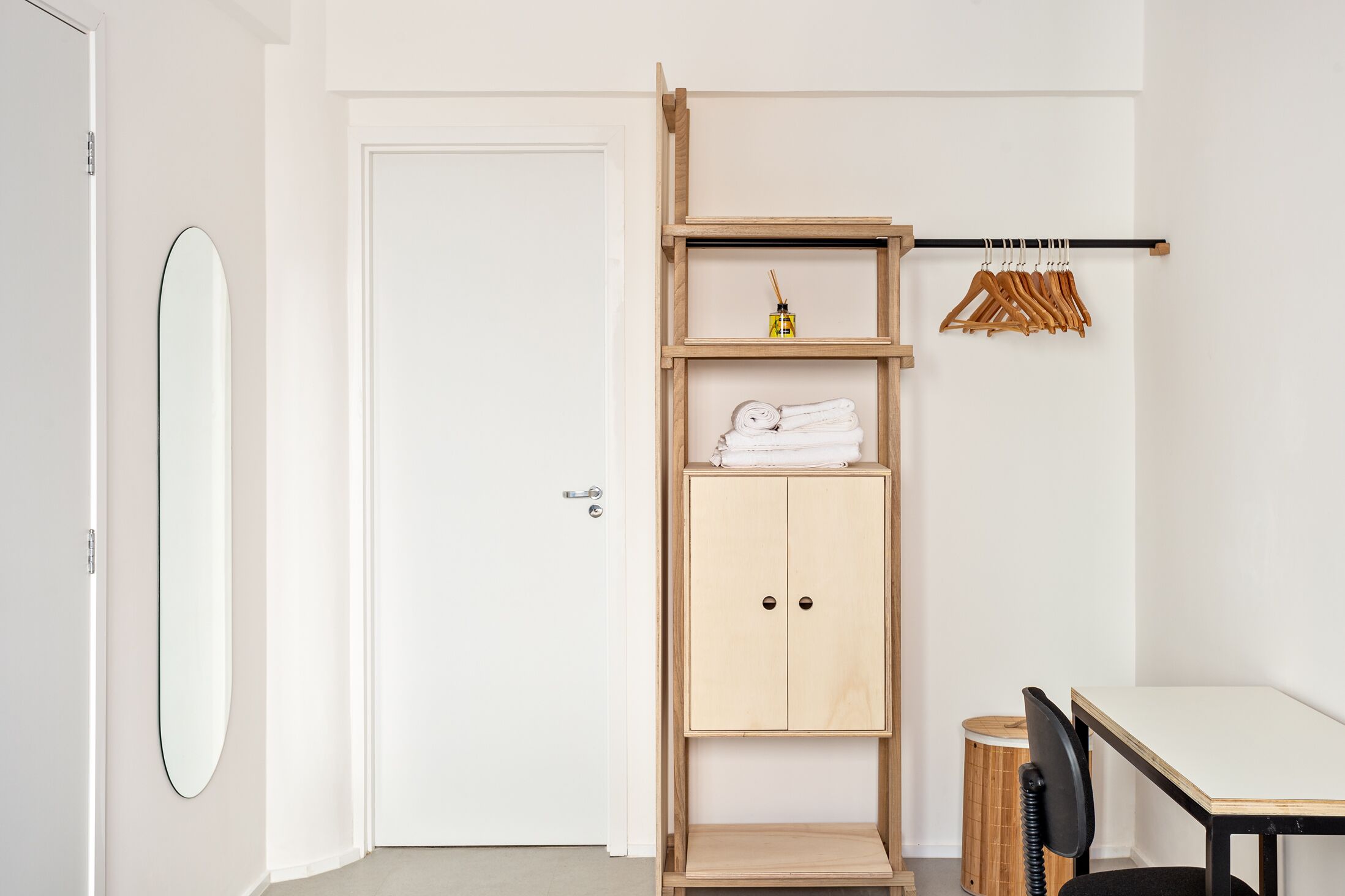 A room with a white door, a wooden shelf unit with a hanging rack, a small stool, and a table.