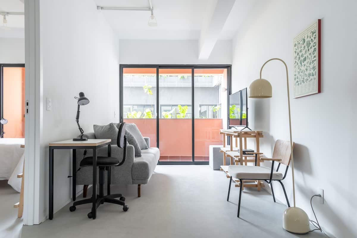 A modern, well-lit room with a desk, office chair, and a sofa. A large window provides a view of the outside.