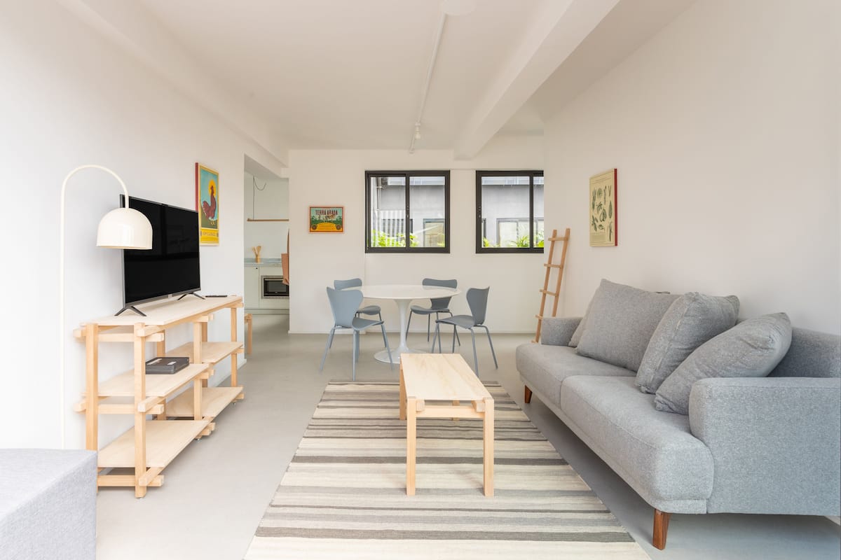 A modern living room with a grey sofa, wooden coffee table, and a TV on a wooden shelf. There is a dining area with a round table and chairs, and a kitchen visible in the background.