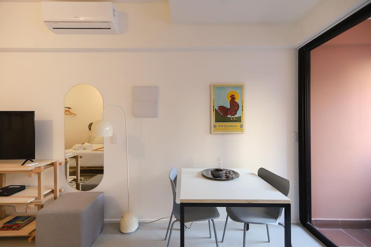A modern, well-lit room with a white table, two chairs, a TV, a bookshelf, and a painting on the wall.