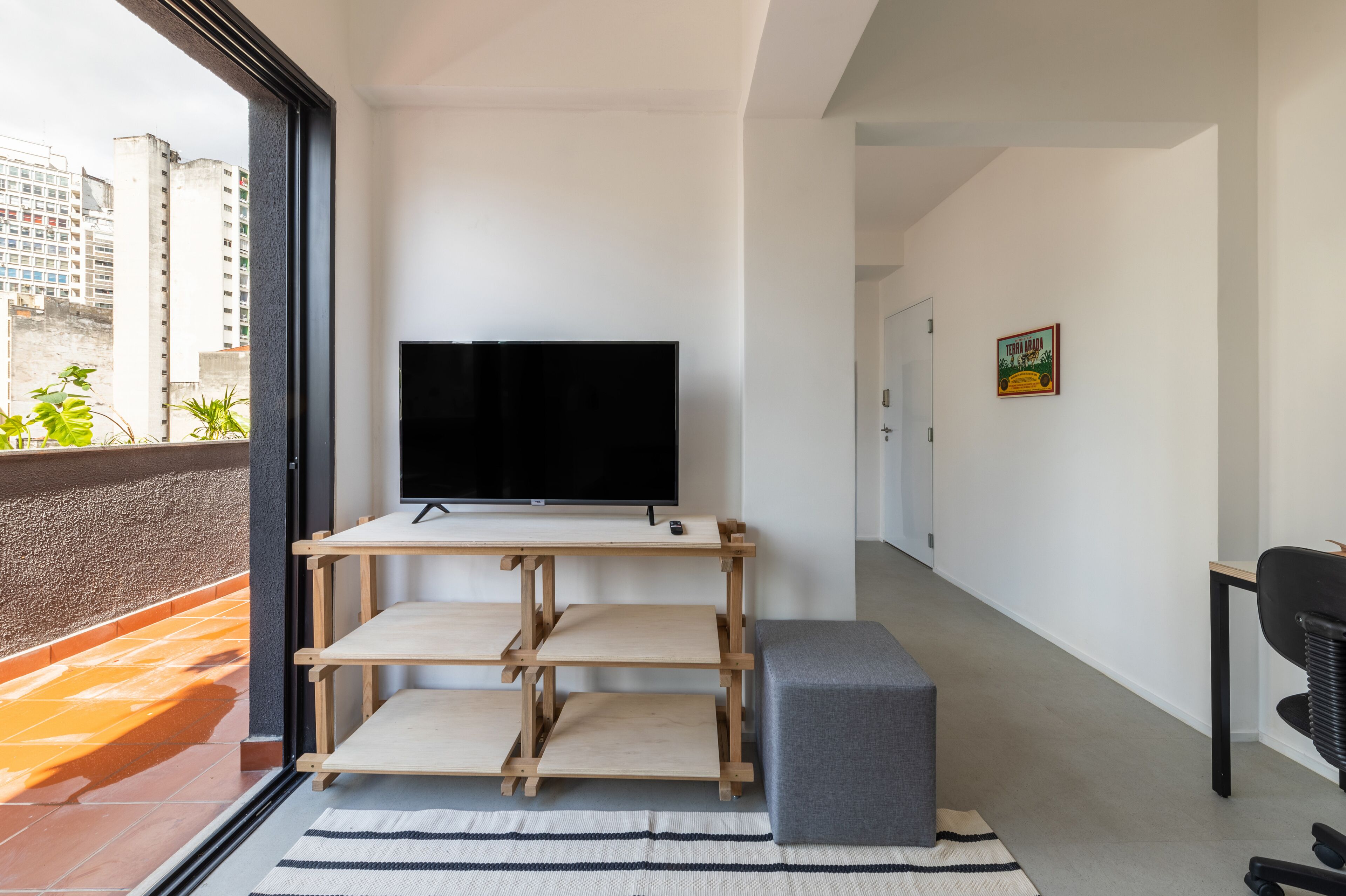A modern living room with a TV on a wooden stand, a grey ottoman, and a view of tall buildings through a balcony.