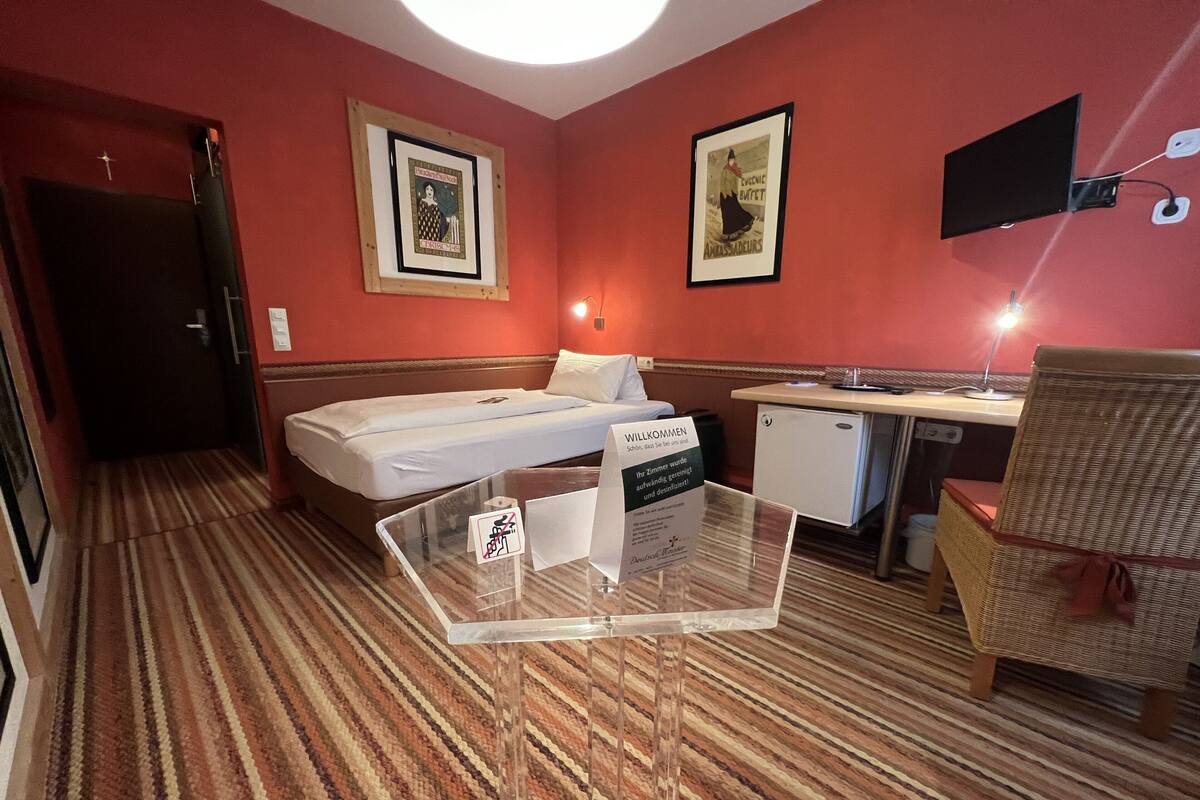A hotel room with a bed, a desk, a chair, a small kitchenette, and a sign indicating the room is ready for guests.
