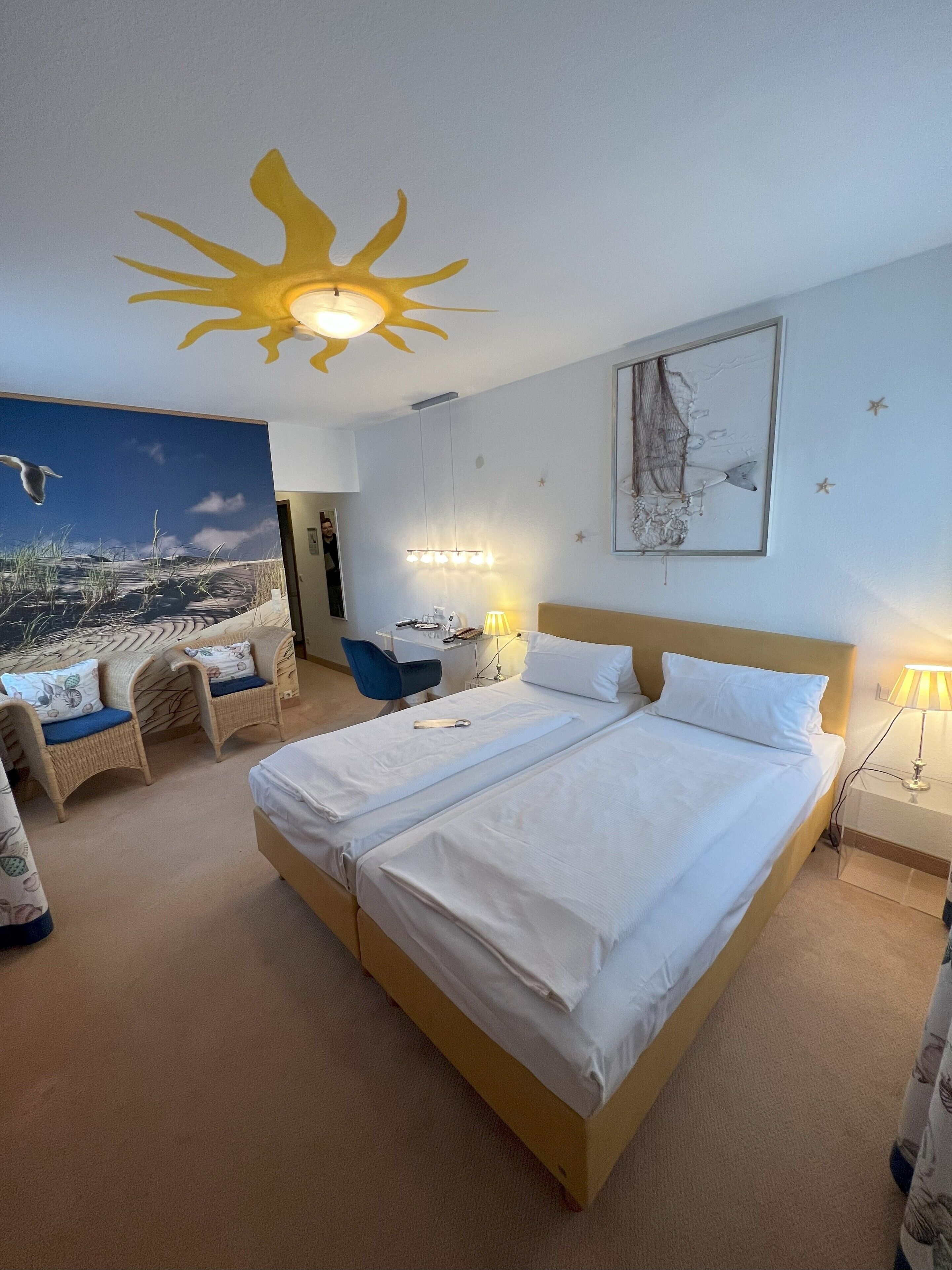 Ein Hotelzimmer mit einem großen Bett, zwei Sesseln, einem Schreibtisch und einem Wandbild mit einer Strandansicht.
