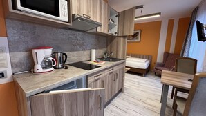 Cocina compacta con microondas, cafetera y hervidor. En la habitación contigua hay una cama y una silla.