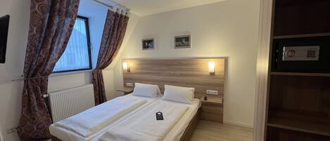 A hotel room with a double bed, a bedside table with a lamp, a wooden headboard, a small shelf, and a window with patterned curtains.
