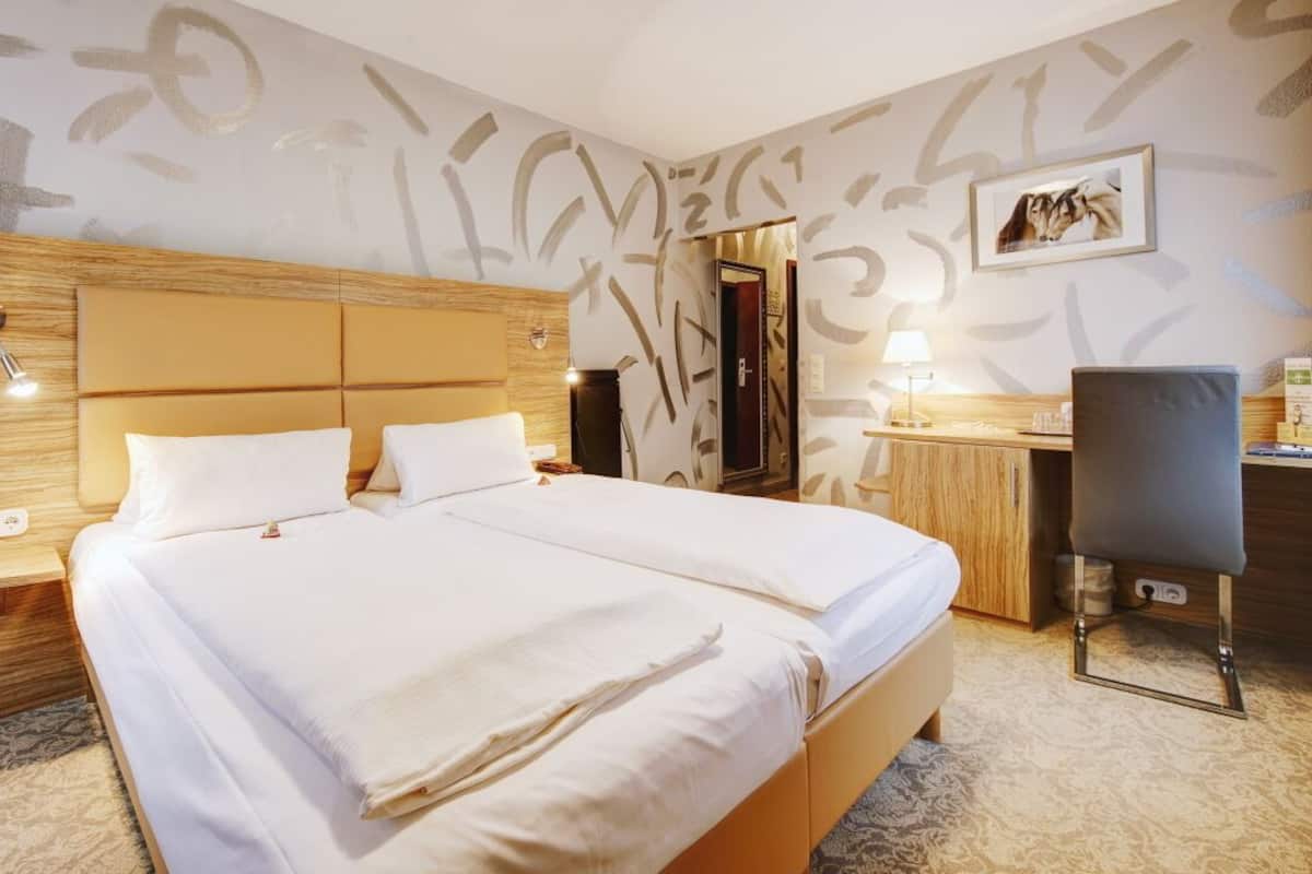 A hotel room with a large bed, wooden headboard, bedside tables, a desk with a chair, and a framed picture on the wall.