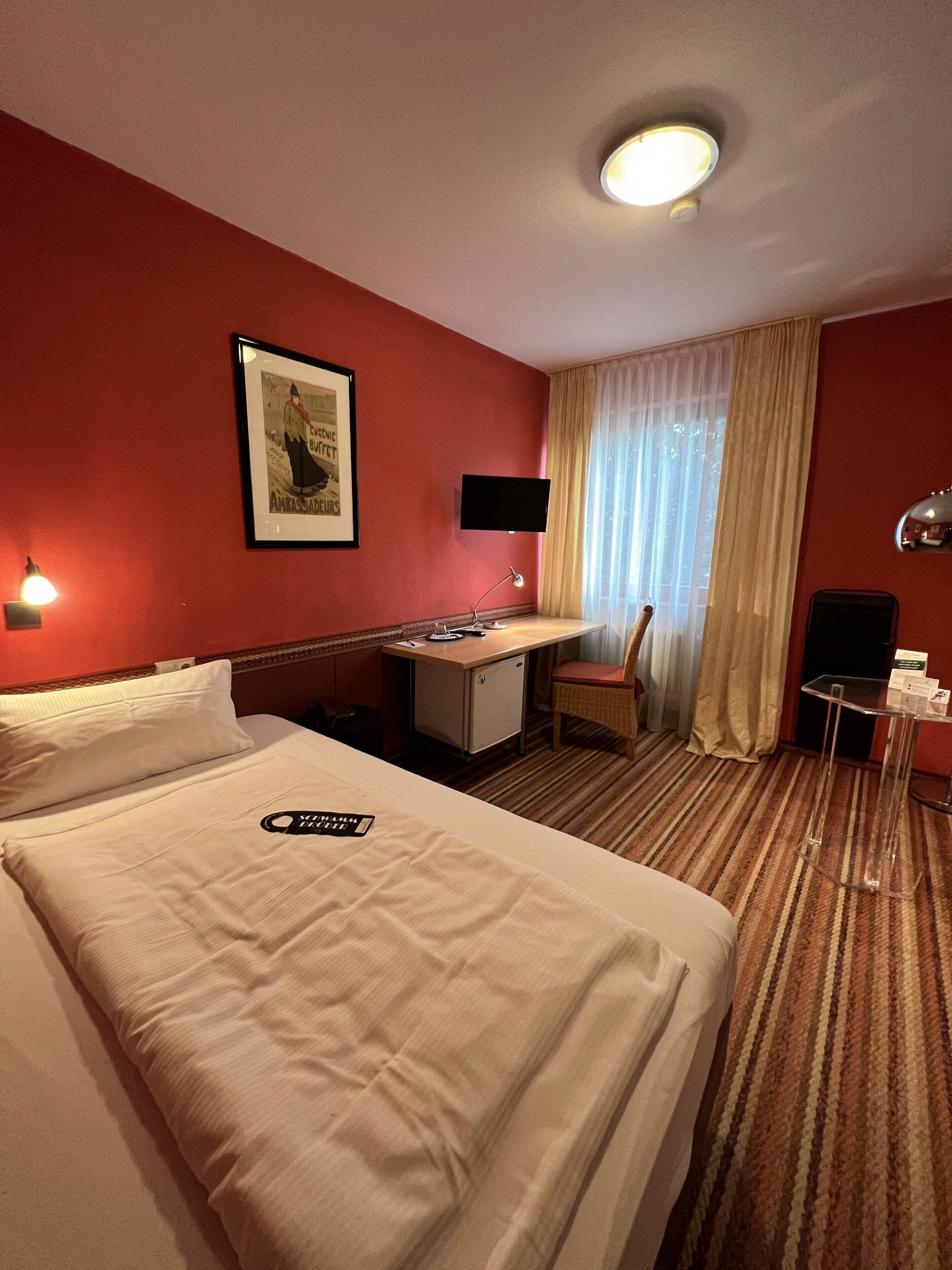 Quarto de hotel com uma cama grande, uma escrivaninha, uma cadeira, uma televisão e uma mesinha.