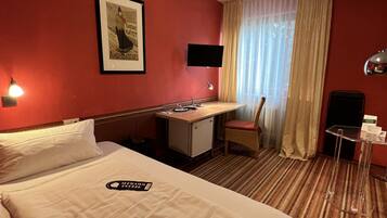 Quarto de hotel com uma cama grande, uma escrivaninha, uma cadeira, uma televisão e uma mesinha.