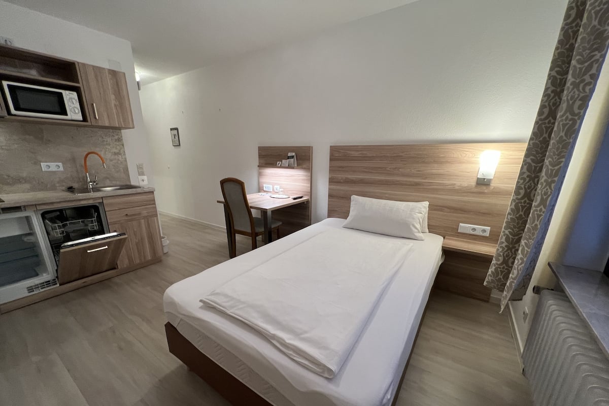 A hotel room with a bed, a desk, a chair, a microwave, and a small kitchenette.