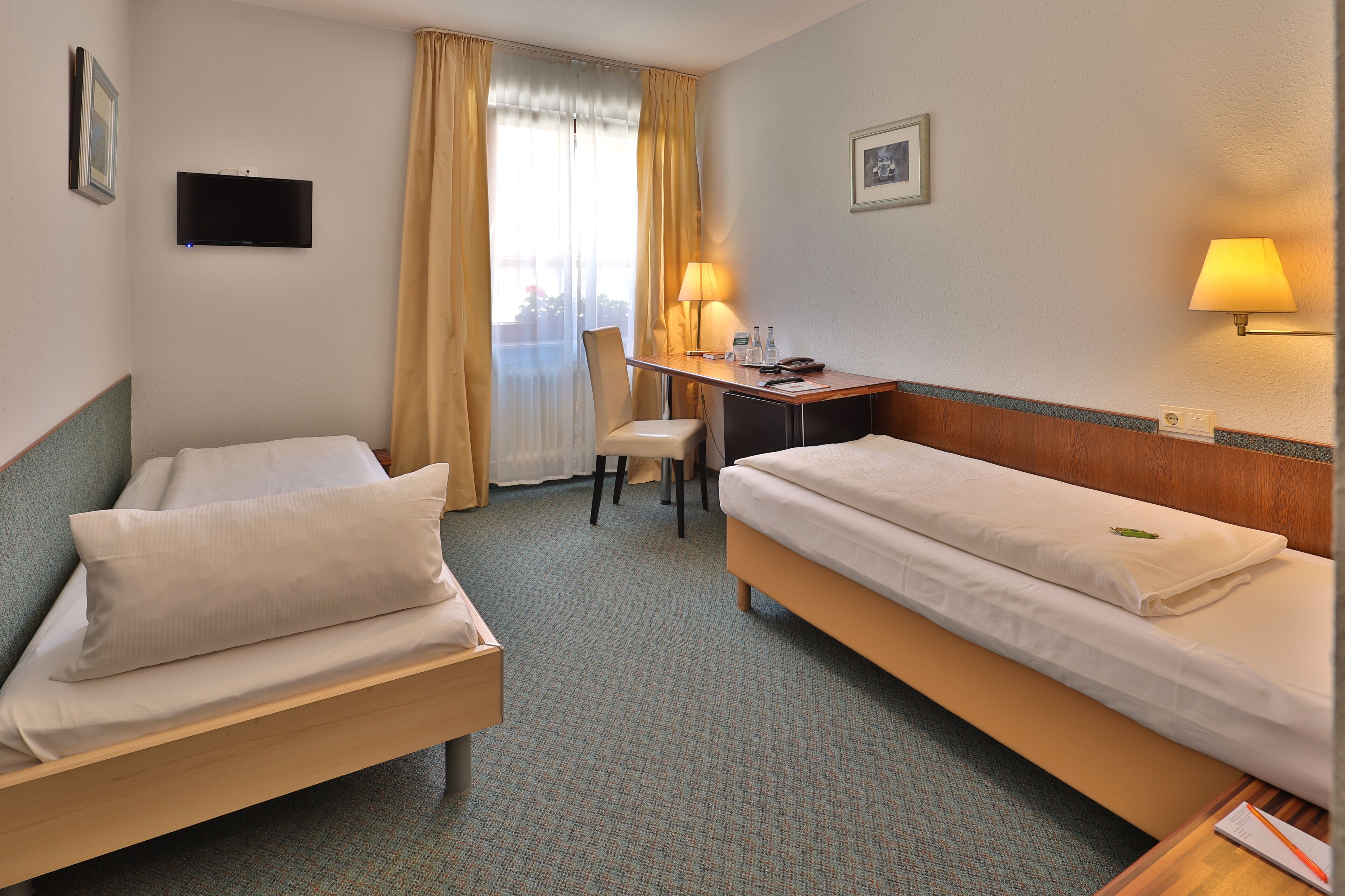 Habitación de hotel con dos camas individuales, un escritorio con silla, un televisor en la pared y una ventana con cortinas.