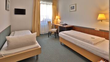 Habitación de hotel con dos camas individuales, un escritorio con silla, un televisor en la pared y una ventana con cortinas.