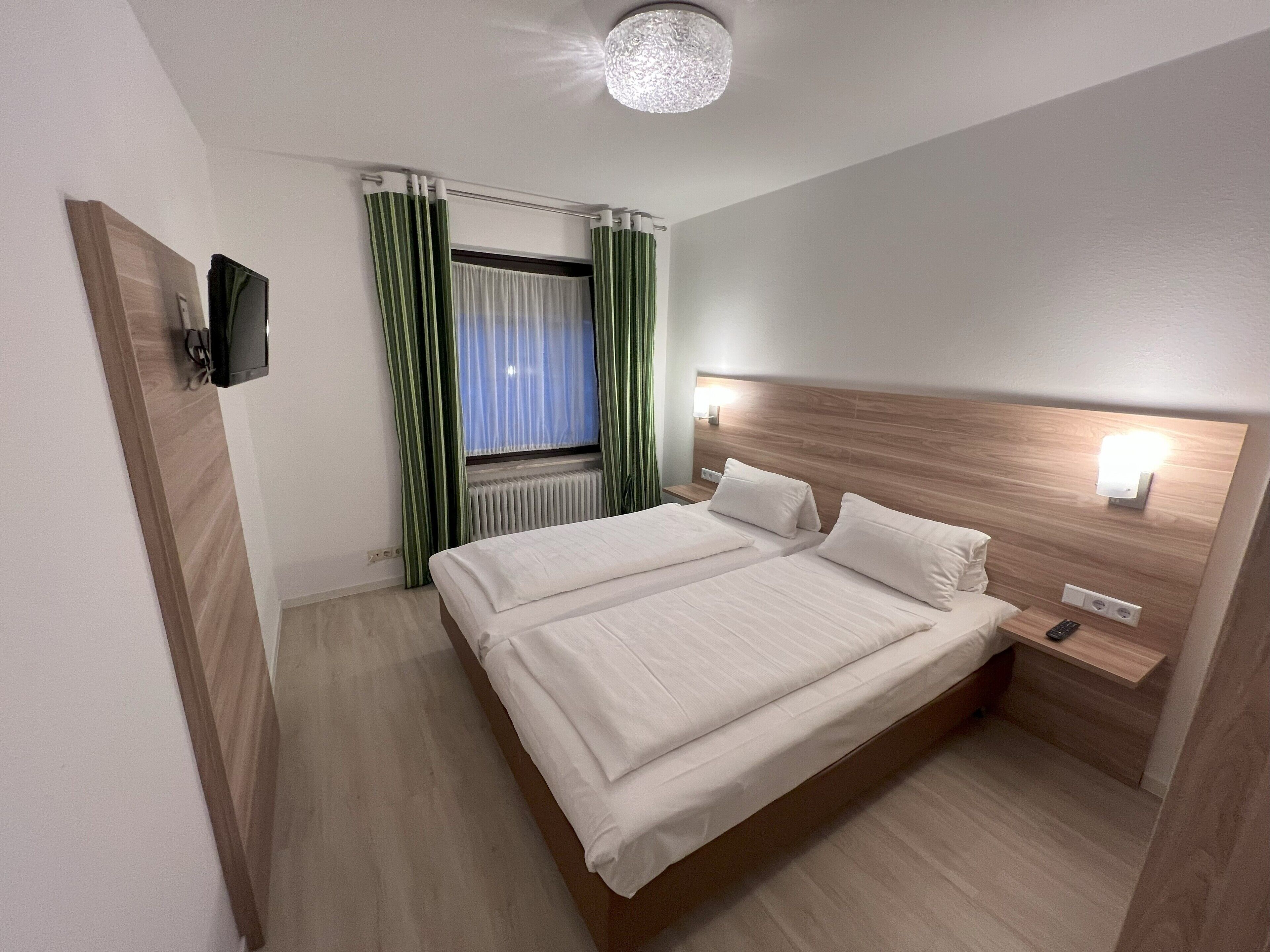 A hotel room with a double bed, a bedside table with a lamp, a TV mounted on the wall, and a window with curtains.