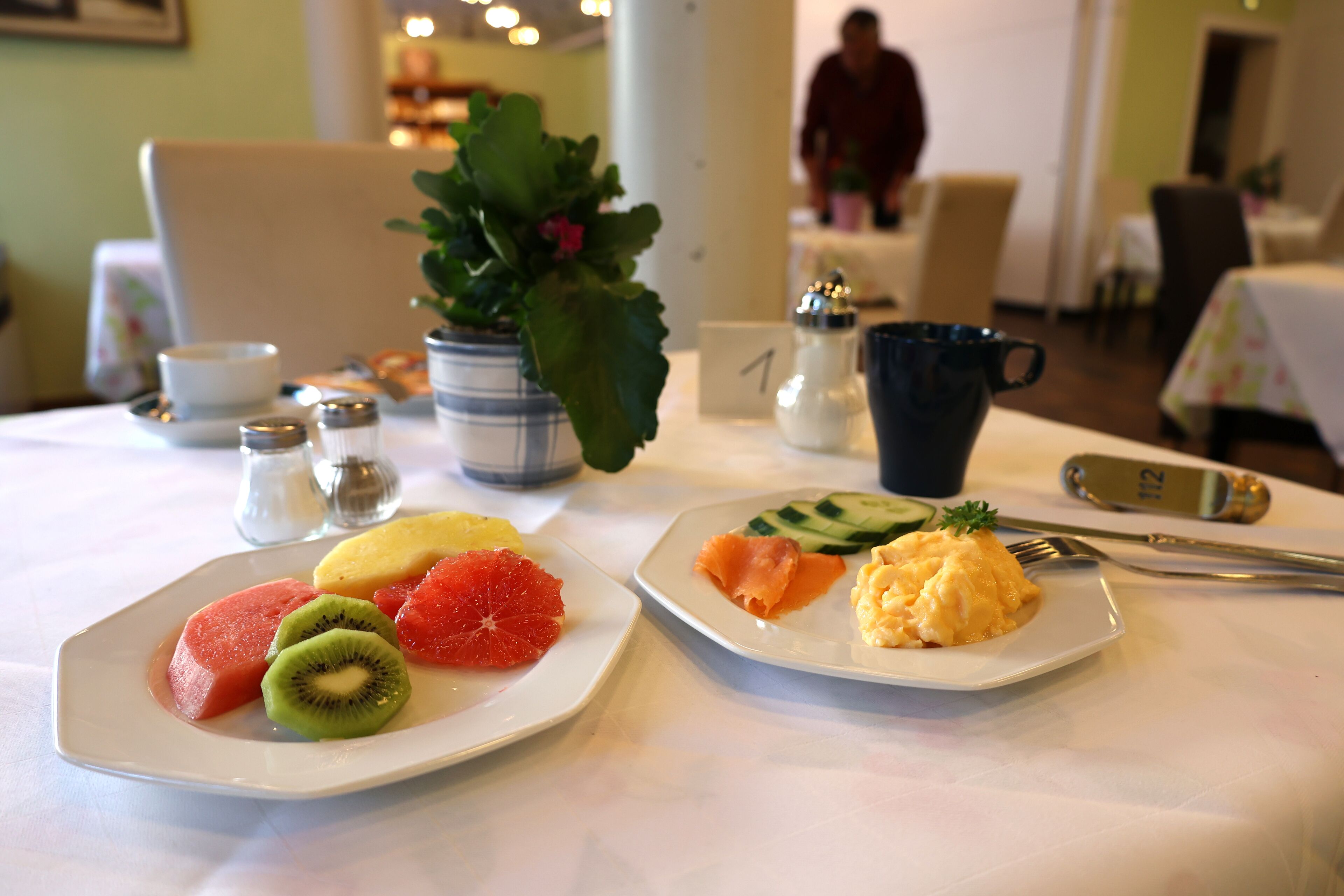 A dining table with plates of fruit and scrambled eggs, salt and pepper shakers, a small potted plant, and a coffee cup.