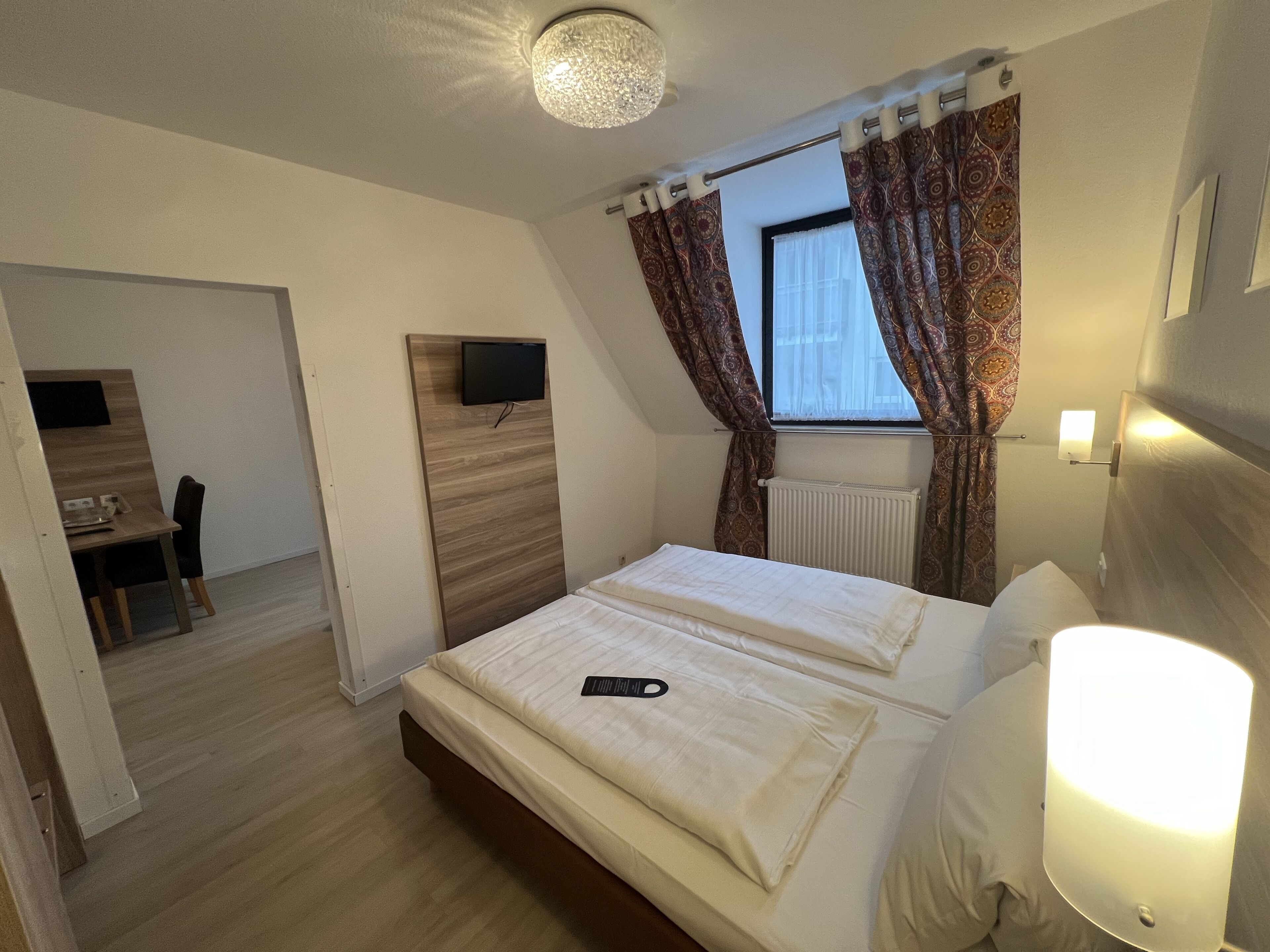 Una habitación de hotel con cama, escritorio, televisor y ventana con cortinas.