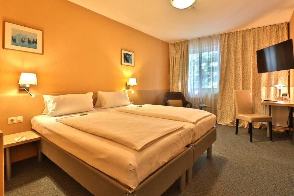 A hotel room with a large bed, two bedside lamps, a desk with a chair, a TV, and two framed pictures on the wall.