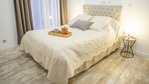 Uma cama bem arrumada com colcha bege, uma mesa de cabeceira com abajur e uma bandeja de vime com bule e xícara.