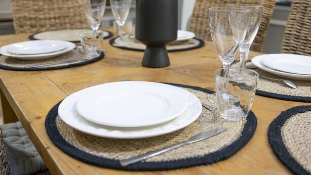 Uma mesa de jantar de madeira posta com pratos brancos, copos transparentes e um vaso de planta.