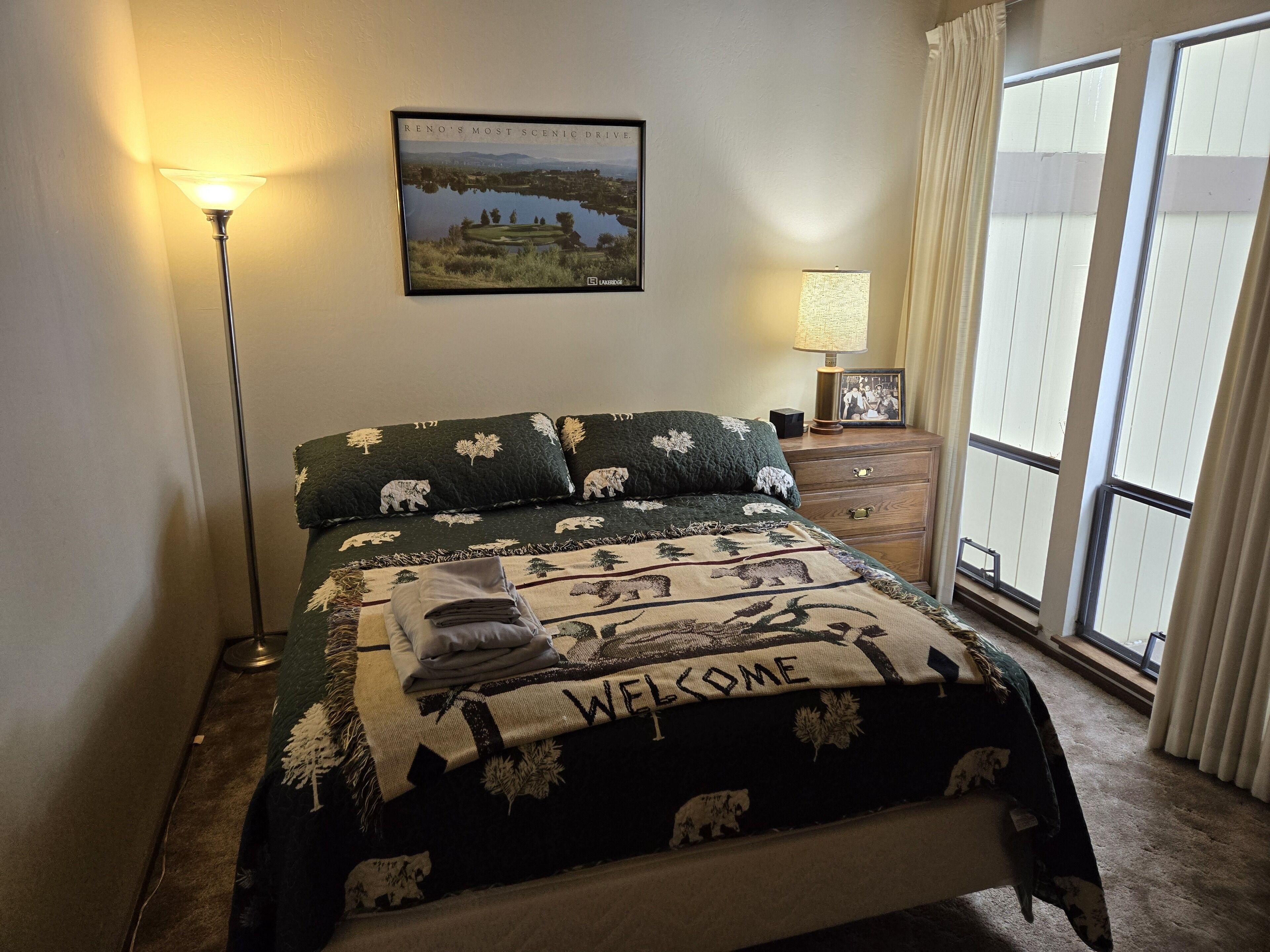 Ein ordentlich bezogenes Bett, ein 'WELCOME'-Schild, ein gerahmtes Panoramafoto an der Wand und eine Lampe auf dem Nachttisch.