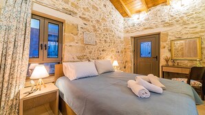 A bedroom with a stone wall, wooden ceiling, a bed with a blue quilt, two bedside lamps, a wooden nightstand, a desk with a mirror, and a door with a decorative glass panel. - Akalli Villa Private Pool & Hot Tub (Paraskevi)