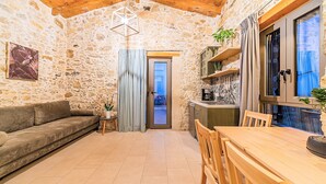 A cozy room with a stone wall, a wooden ceiling, a sofa, a dining table with chairs, a small kitchen area, and a window with curtains. - Akalli Villa Private Pool & Hot Tub (Paraskevi)