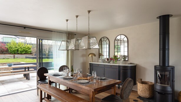 A dining area with a wooden table set for four, a black stove, and a large window with a view of the outdoors. - La perle de mer - Charmante maison avec jacuzzi (Erquy)