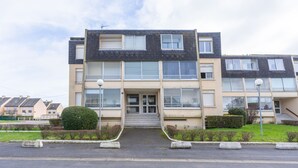 A multi-story residential building with a central entrance, large windows, and a small garden area with hedges.