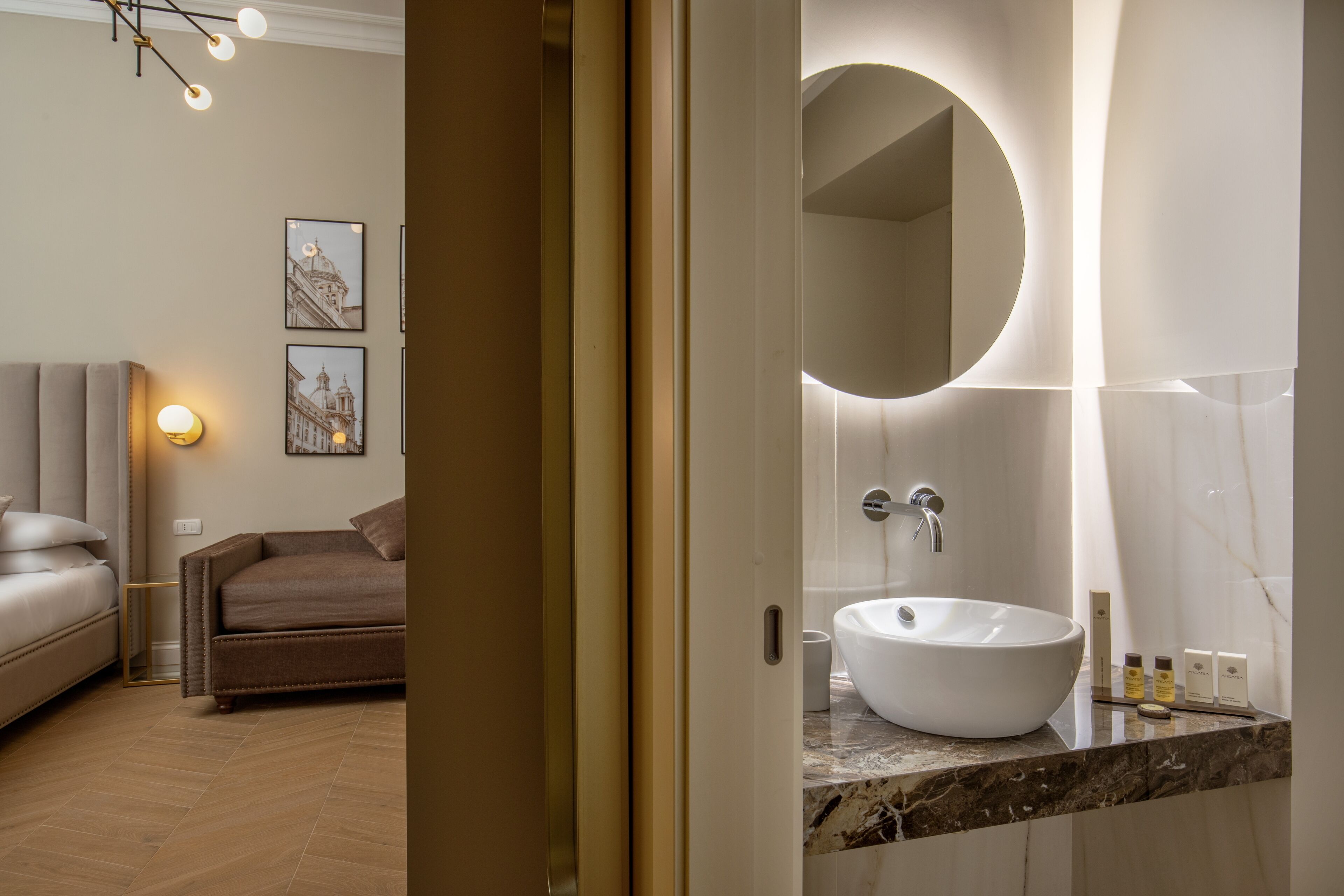 Une salle de bain moderne avec un grand miroir rond, un lavabo blanc et un comptoir en marbre. On y trouve également un canapé marron et un tableau encadré accroché au mur.