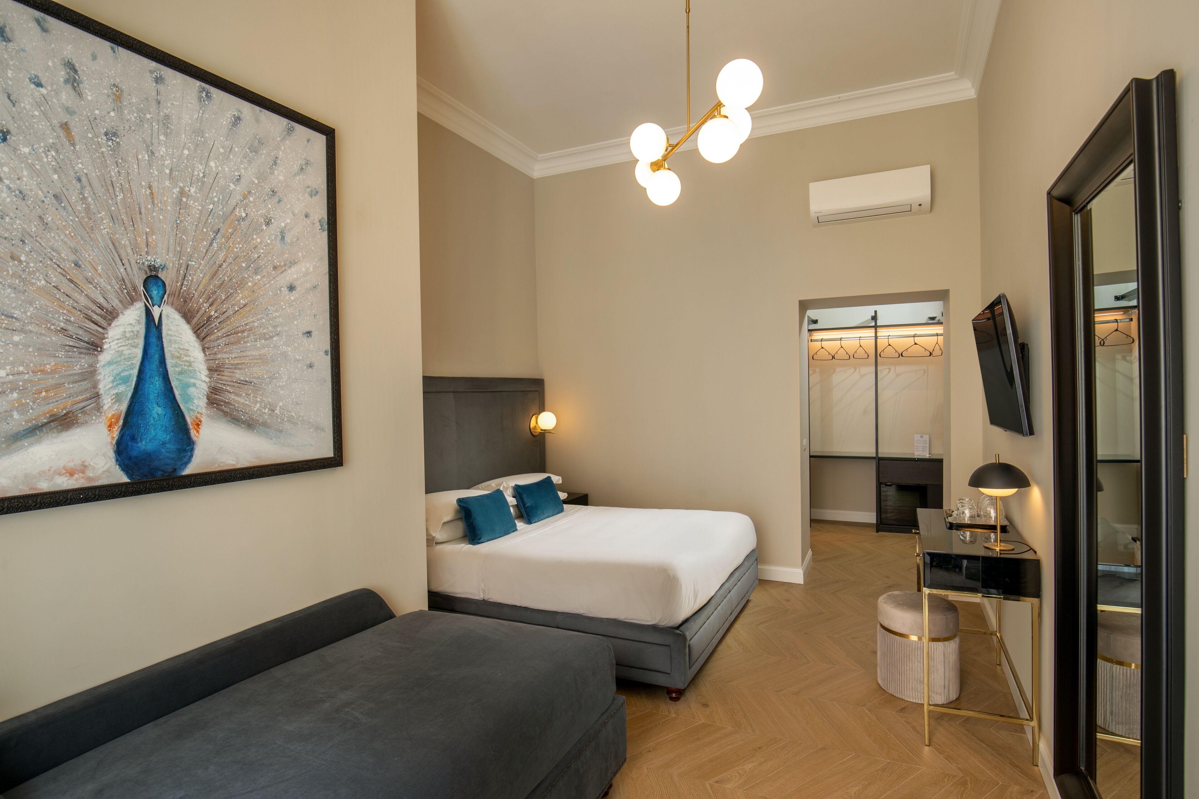 Una habitació d'hotel moderna amb un gran quadre de pavó, un llit amb roba blanca i almoinos blaus, un sofà gris, un escriptori amb una làmpada i un armari amb mirall.