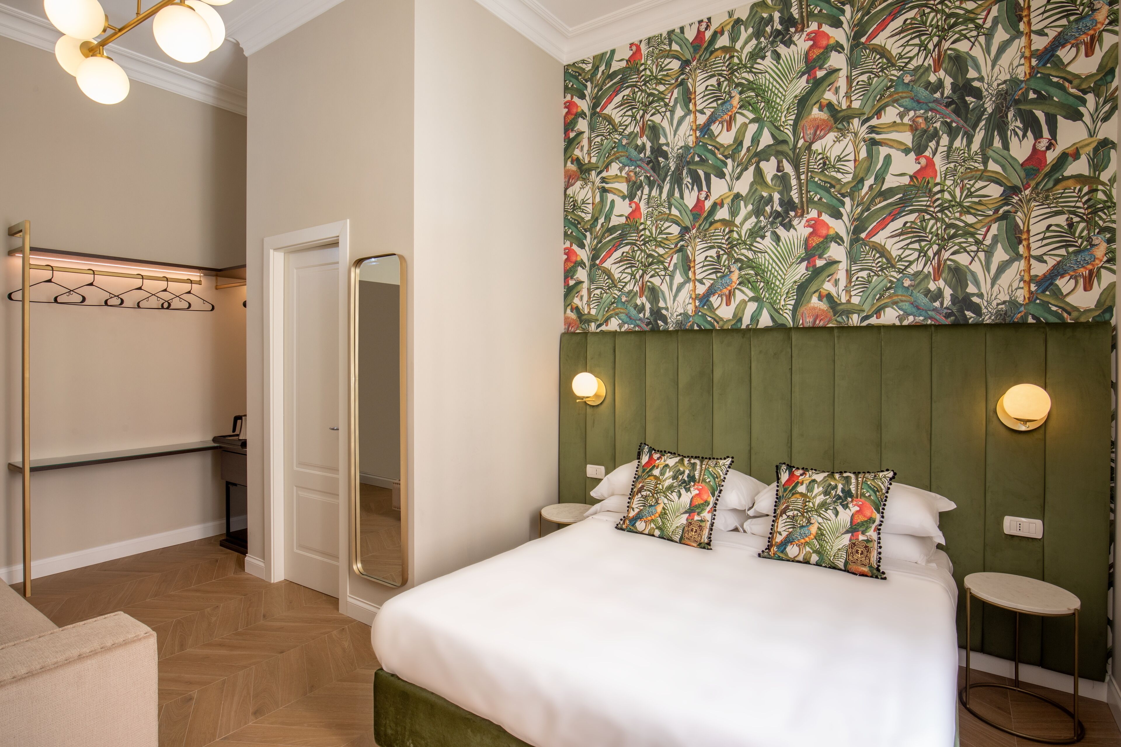 A hotel room with a bed, a green headboard, a wall with a tropical wallpaper, a mirror, and a small round table.