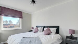 Cama bem arrumada com lençóis brancos e duas almofadas roxas, mesa de cabeceira com abajur e janela com vista para prédios.