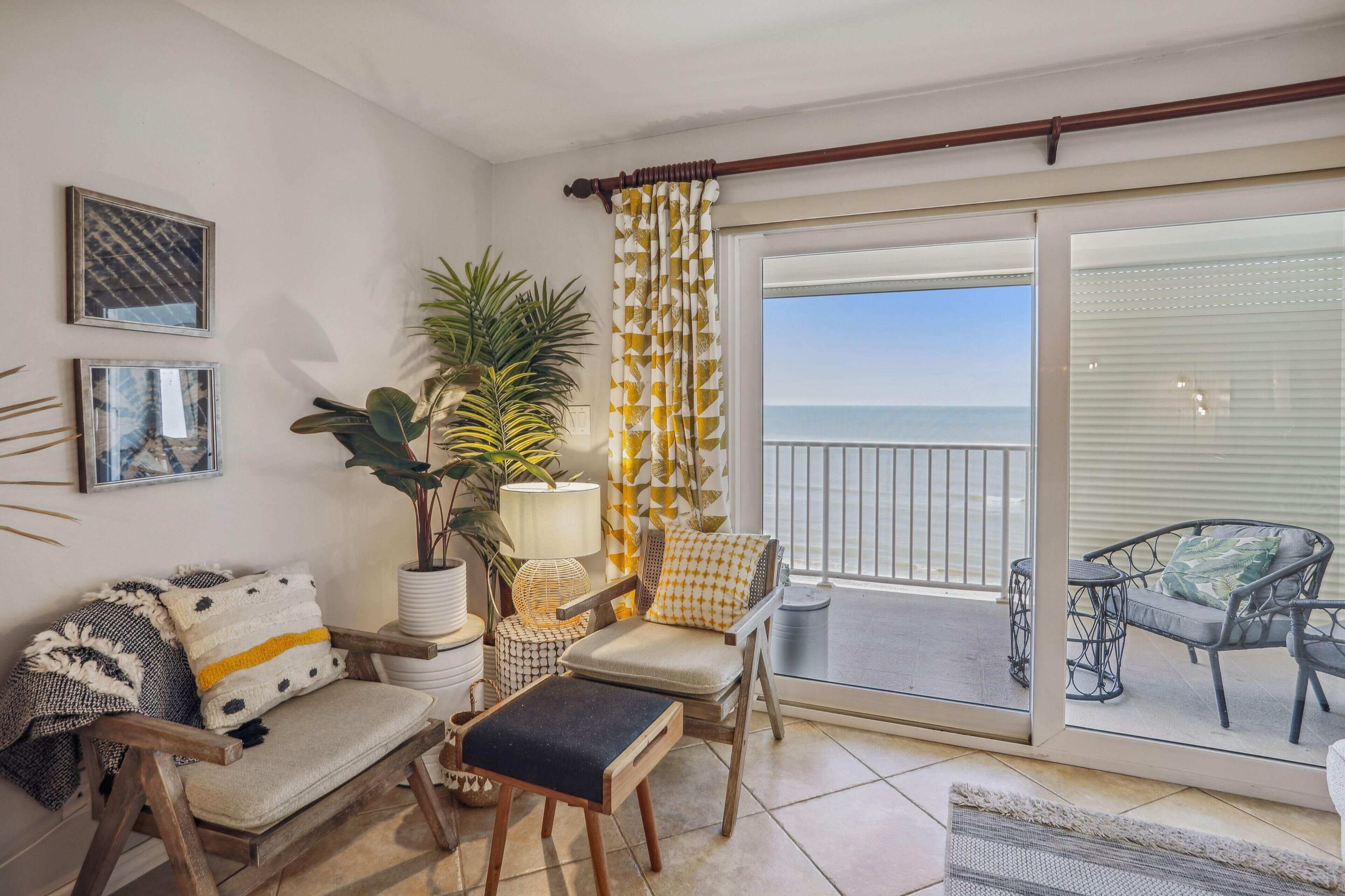 A cozy living area with a sofa, armchair, and ottoman, a potted plant, and a view of the sea through a sliding glass door.