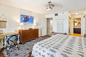 A bedroom with a large bed, a desk, a chair, a dresser, a wall-mounted TV, and a door leading to another room. - Arena De Madeira 502- Premier (Madeira Beach)