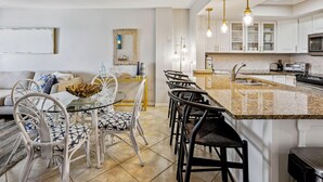 A modern kitchen with a granite island, white cabinets, and stainless steel appliances. There is a dining area with a glass table and metal chairs. - Arena De Madeira 502- Premier (Madeira Beach)