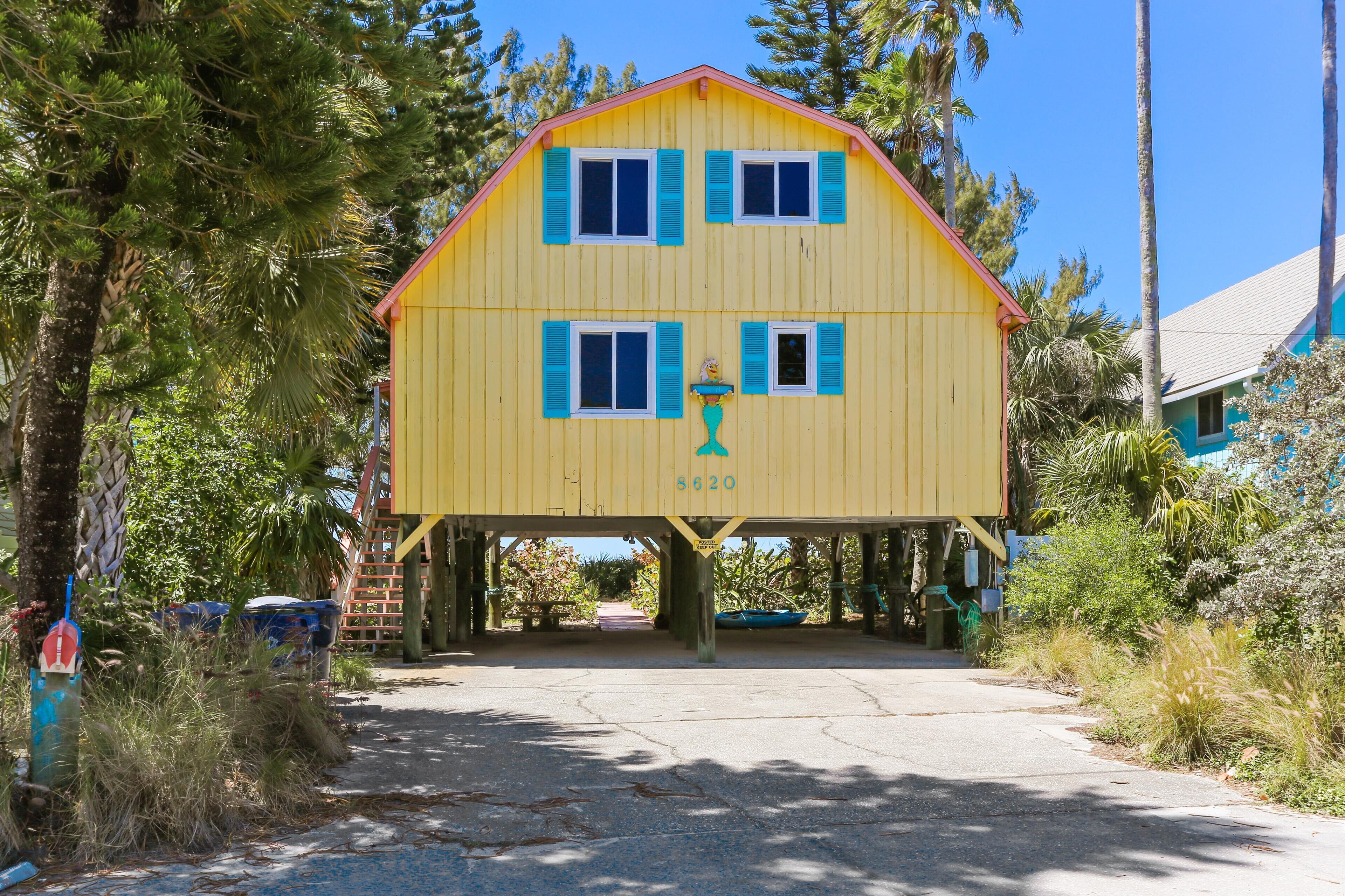 A two-story yellow house with blue shutters, a porch, and a small boat under the house.