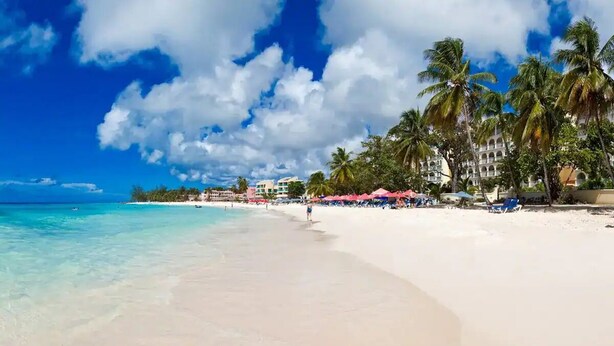 A beach with clear turquoise water, white sand, and palm trees. There are buildings with balconies and umbrellas in the background. - Safe & Spacious 2 BR Apt, Budget Friendly on South
 (Oistins)