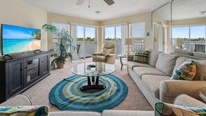 A living room with a television, a ceiling fan, a sofa, a coffee table, and a view of the beach. - Amadeus 9 (Treasure Island)