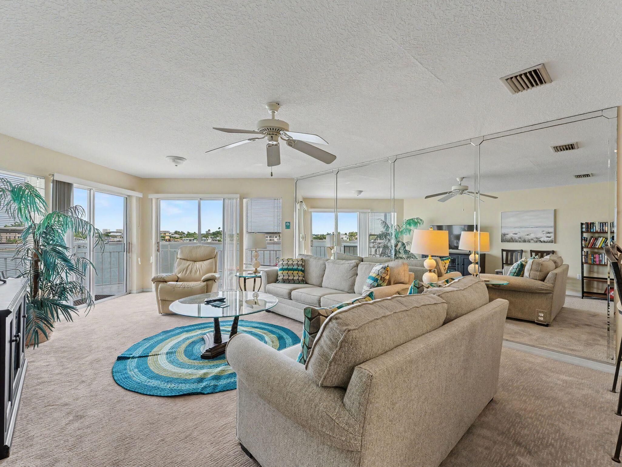 A spacious living room with a sofa, armchairs, a coffee table, and a TV. There are ceiling fans, a bookshelf, and a large window with a view.