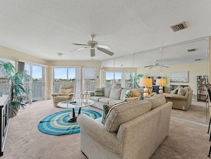 A spacious living room with a sofa, armchairs, a coffee table, and a TV. There are ceiling fans, a bookshelf, and a large window with a view. - Amadeus 9 (Treasure Island)