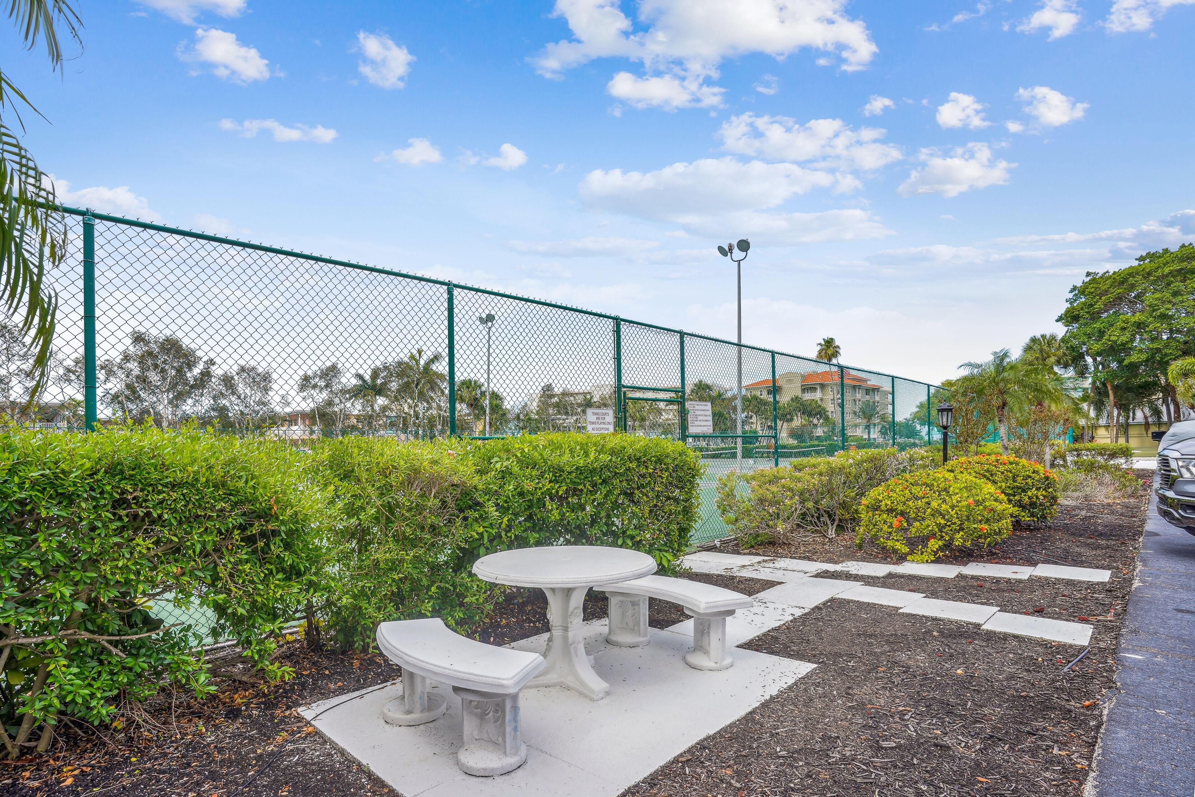 A fenced outdoor area with a white table and benches, surrounded by greenery and a clear sky.
