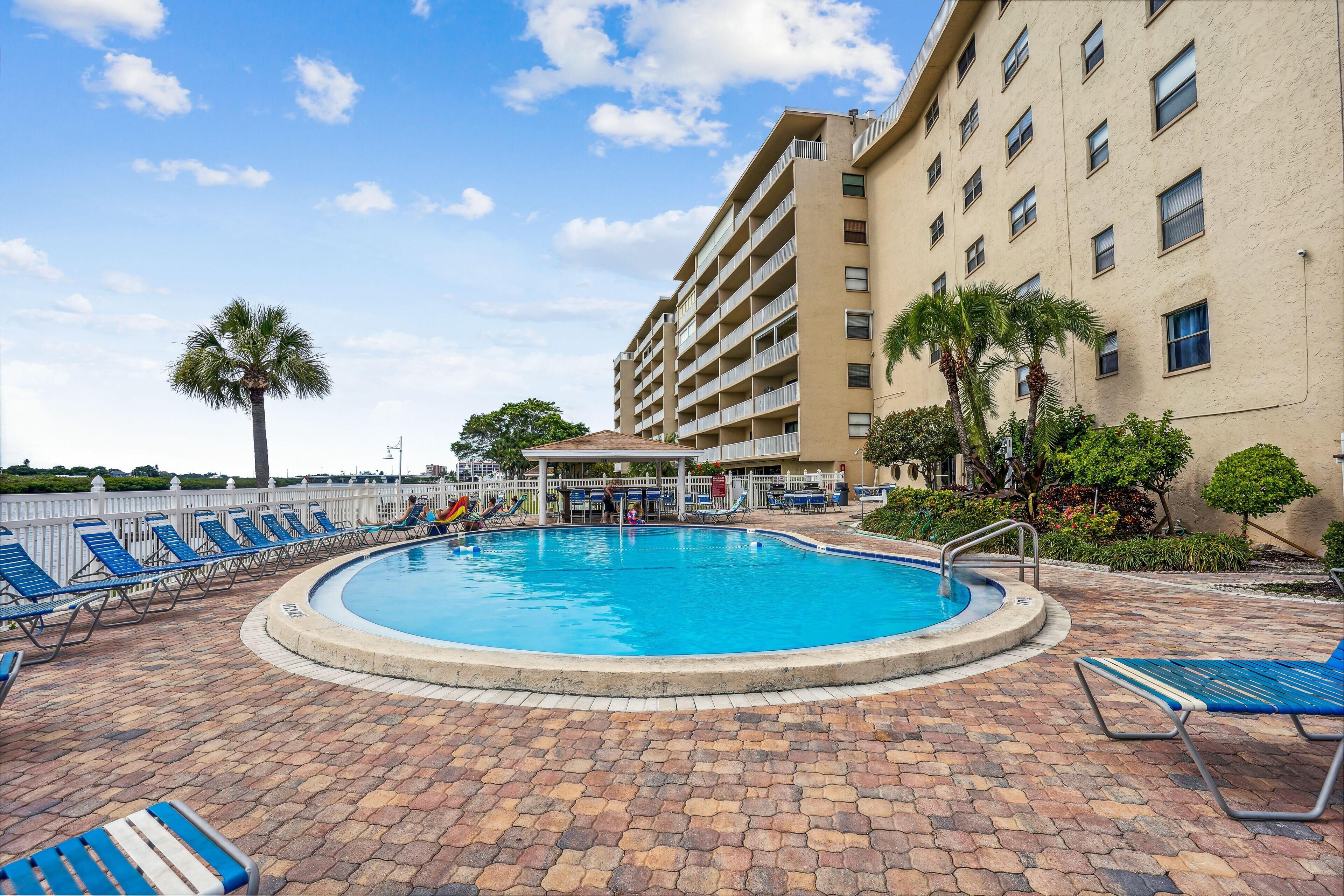 A swimming pool with lounge chairs, a covered seating area, and a building with multiple balconies.