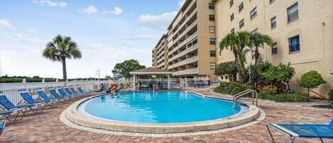 A swimming pool with lounge chairs, a covered seating area, and a building with multiple balconies.