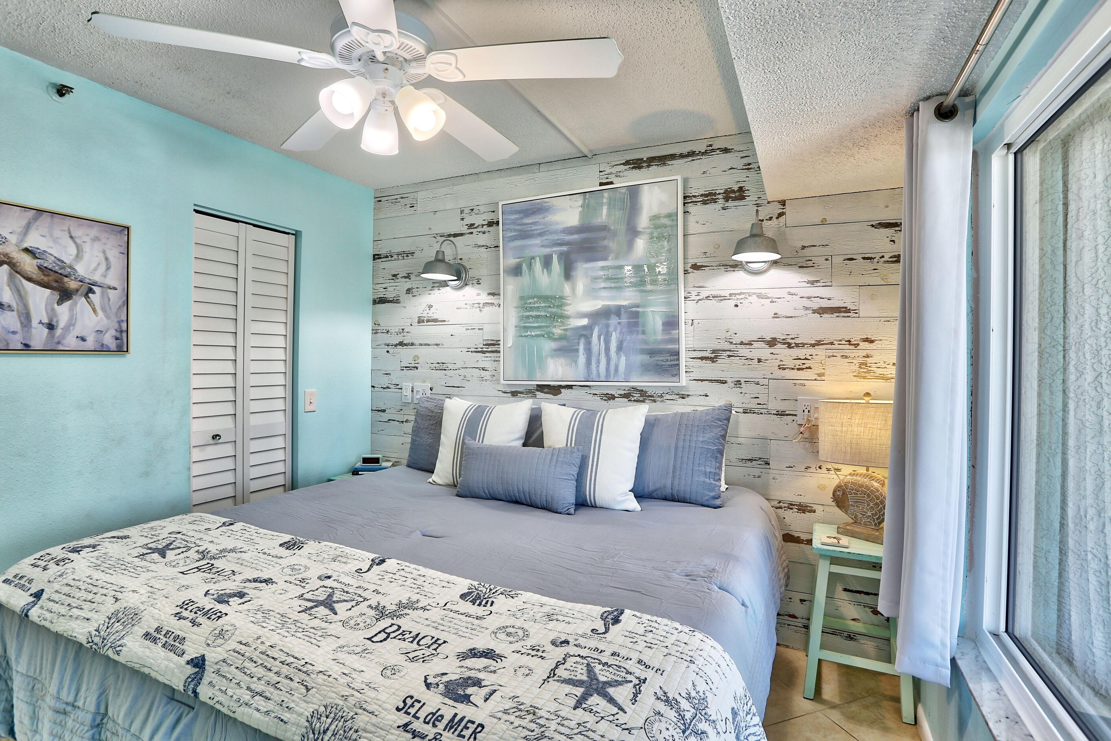 A bedroom with a bed, a ceiling fan, a window with curtains, and a wall with a distressed wood panel design.