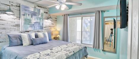 Un dormitorio con cama, ventilador de techo, ventana con cortinas y un espejo que refleja un trípode.